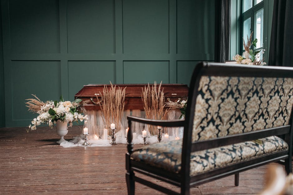 A floral arrangement placed on the floor in front of a wooden funeral casket, featuring dried pampas grass, white roses, and greenery in a white ceramic vase. Surrounding the arrangement are small whi