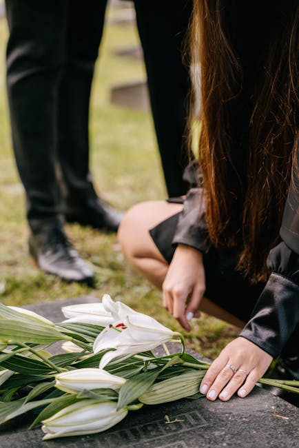 A woman with long brown hair, dressed in dark clothing, kneels on the ground to arrange white lilies with long green leaves on a stone memorial or tablet, likely during a funeral or memorial service.