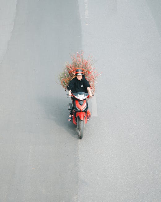 A delivery person wearing a black helmet and black jacket is riding a red scooter on a paved road. Attached to the rear of the scooter is a large, vibrant floral arrangement consisting of pink and ora