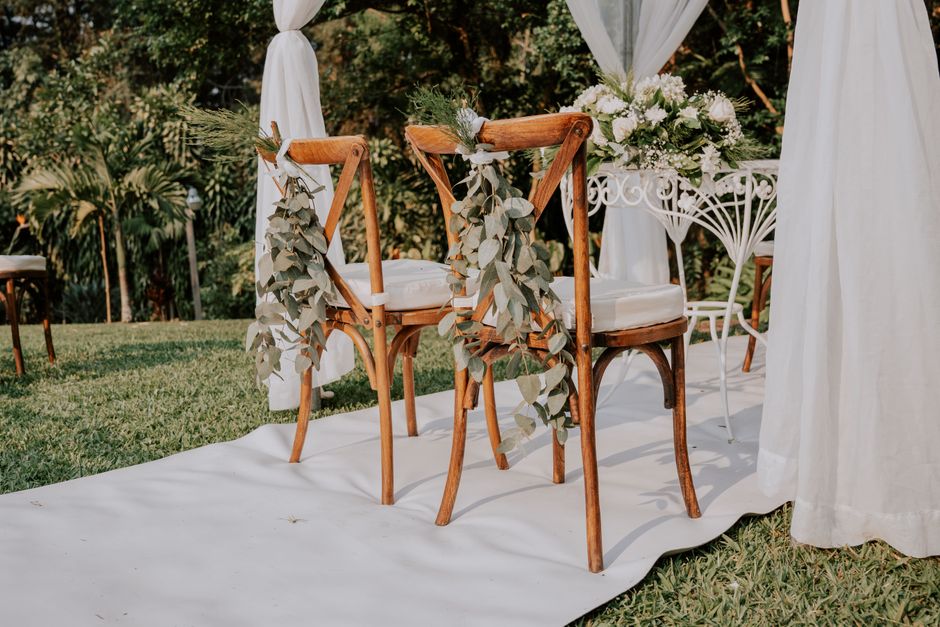 A pair of wooden chairs with beige cushions are decorated with cascading eucalyptus garlands, positioned next to a white floral arrangement on a white ornate metal table at an outdoor wedding ceremony
