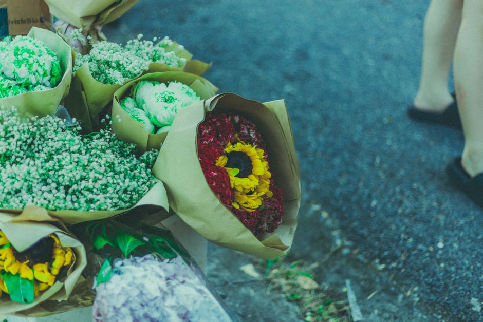 A collection of floral arrangements and bouquets placed on the ground outdoors, featuring various flowers wrapped in brown paper and green cellophane. Prominent blooms include bright yellow flowers, d