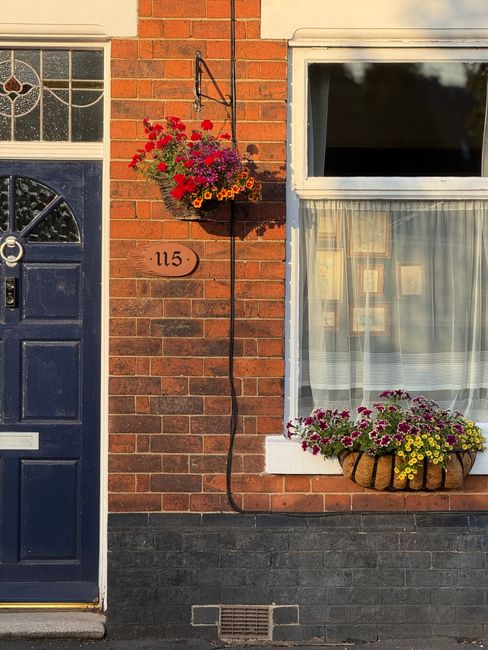 A close-up of a brick house façade featuring a dark blue front door with a silver handle and letterbox, and a large window with white framing and sheer curtains inside. To the left of the door, a han